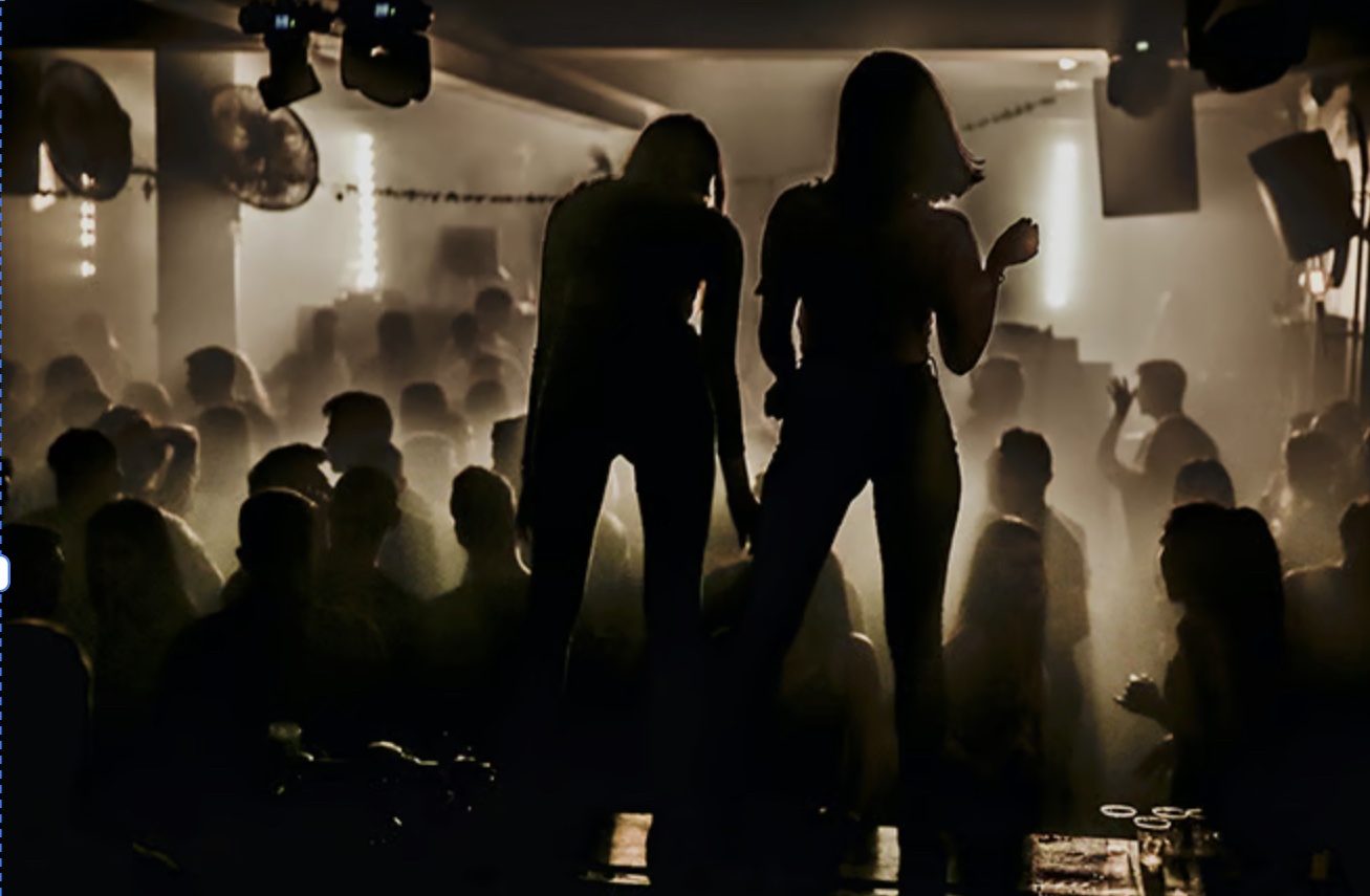 Two silhouetted performers stand on a raised platform above a crowded, dimly lit nightclub dance floor with hazy backlighting and fog.