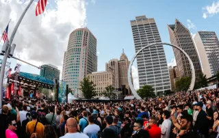 Crowd at an outdoor city concert in a downtown plaza with tall buildings and a circular sculpture nearby