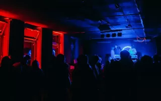People dancing in a dark club with red-lit walls on the left and a blue-lit stage where two DJs perform on a dimly lit platform.