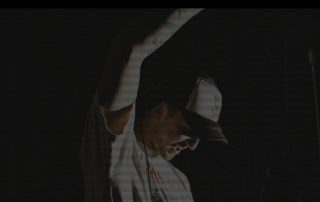 Man in a white shirt and light cap raises his arm on a dark stage, with a flame glow in the background on the right.