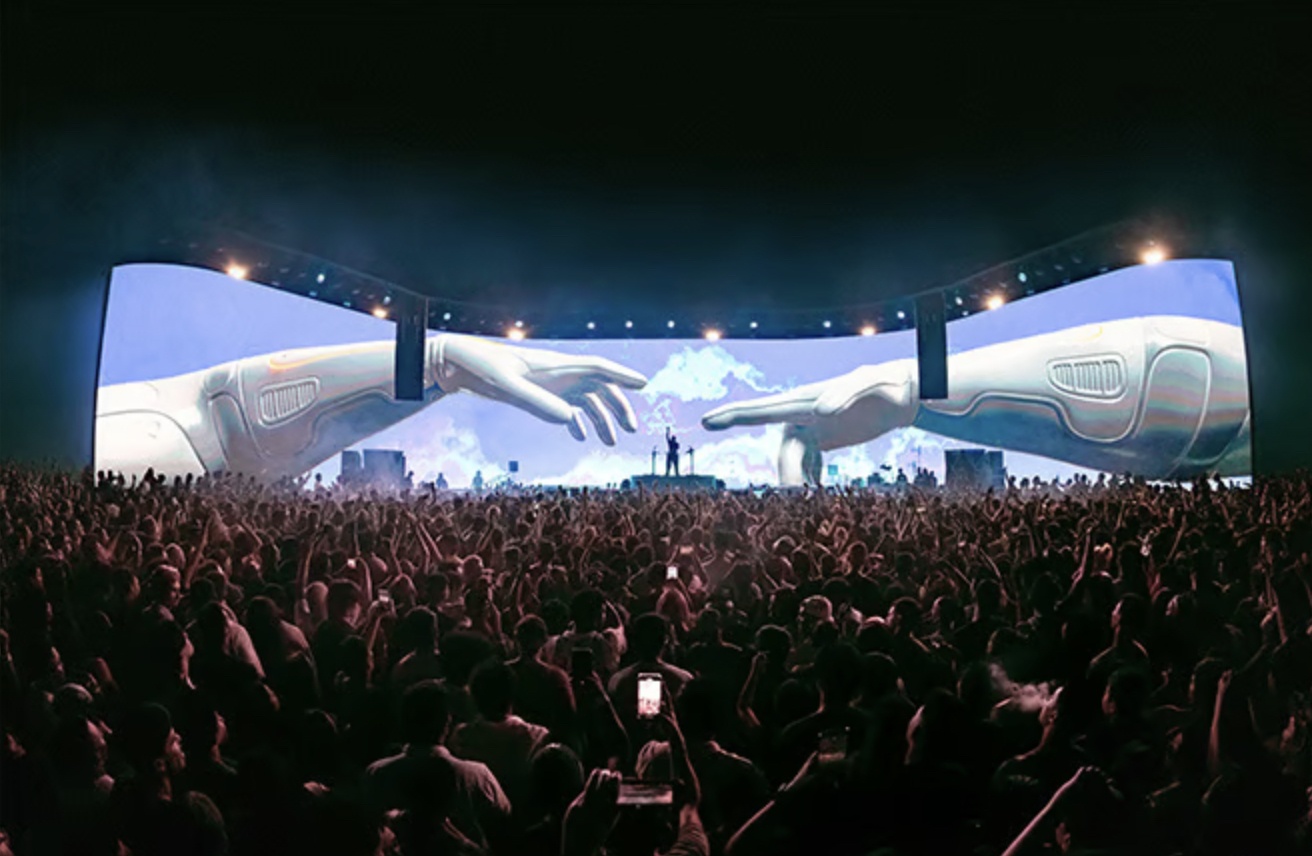 Massive curved stage screens at a concert show two robotic hands reaching toward each other above a packed crowd at night.
