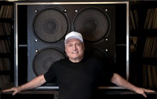 Portrait of a smiling man in a white cap and black shirt, leaning on a large speaker cabinet in a music studio.