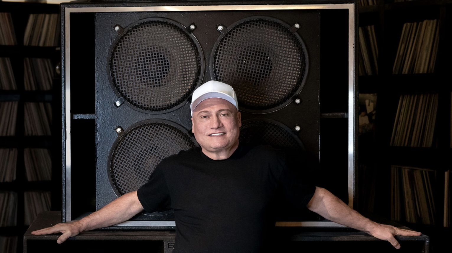 Portrait of a smiling man in a white cap and black shirt, leaning on a large speaker cabinet in a music studio.