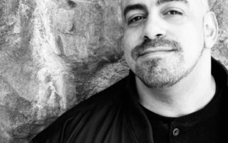 Black-and-white portrait of a bald man with a short beard smiling slightly, against a rough stone wall backdrop, wearing a dark jacket.