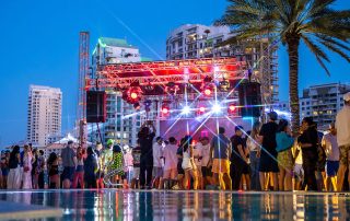 Crowd gathered around a lit outdoor stage beside a swimming pool at night, with palm trees and tall buildings in the background.