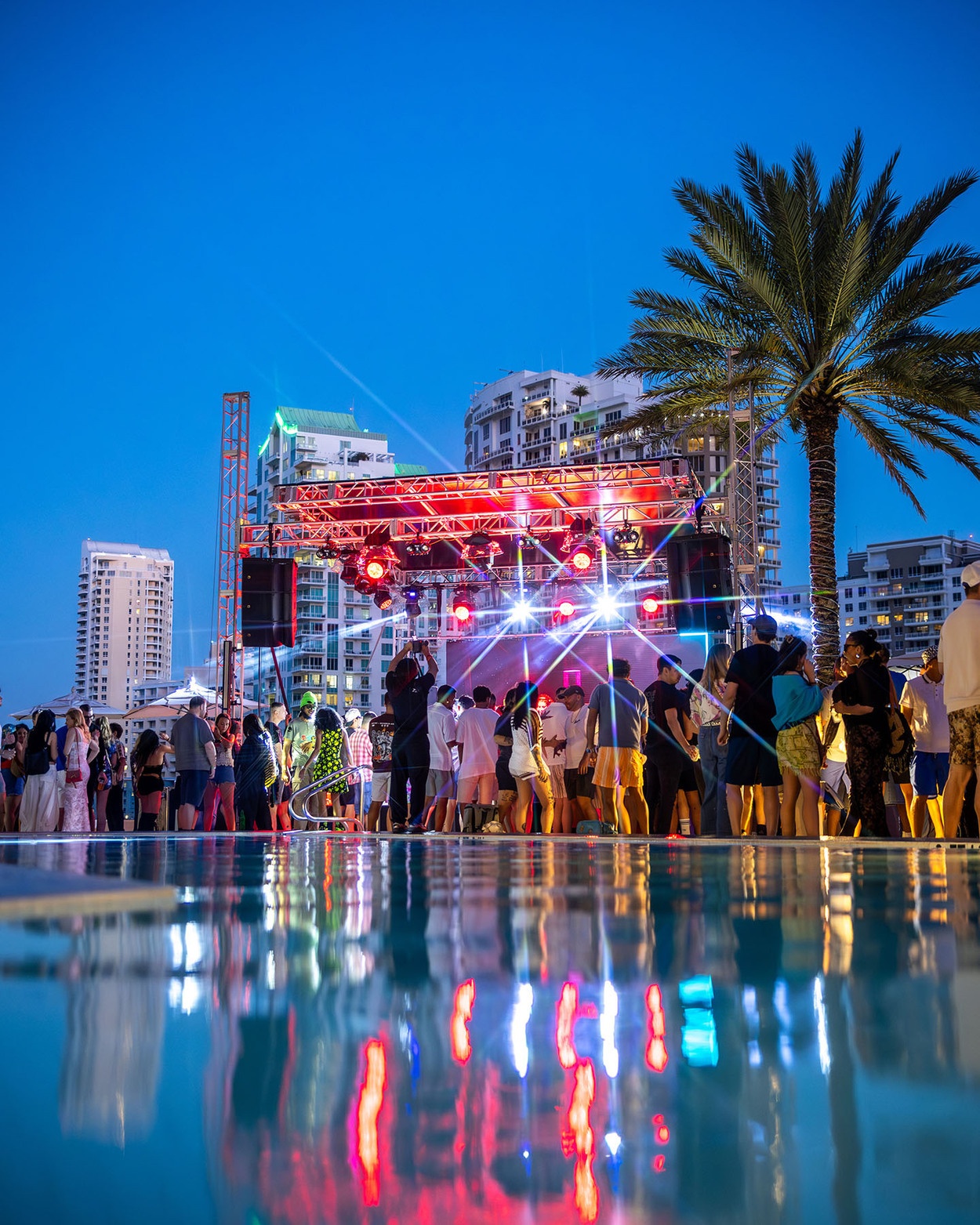 Crowd gathered around a lit outdoor stage beside a swimming pool at night, with palm trees and tall buildings in the background.