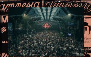Large indoor concert venue filled with a dense crowd facing a brightly lit stage with truss lighting rigging above.