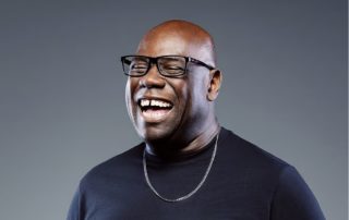 Portrait of a joyful Black man wearing glasses and a dark shirt, laughing with mouth open against a gray backdrop.