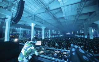 DJ performing at a large indoor concert with a packed crowd and blue stage lighting.