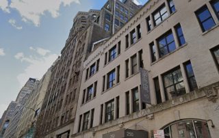 Tall city street with beige brick building and large windows, storefront at 250 on the ground level, banner hanging on the facade.