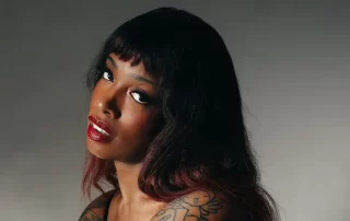 Portrait of a woman with dark hair and red lipstick, looking toward the camera, with a shoulder tattoo visible in a studio setting.