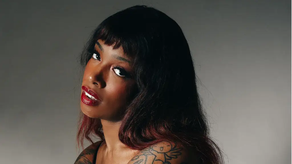 Portrait of a woman with dark hair and red lipstick, looking toward the camera, with a shoulder tattoo visible in a studio setting.
