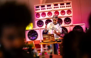 DJ performing on stage with a laptop, colorful giant speaker stacks behind him, colorful club lighting