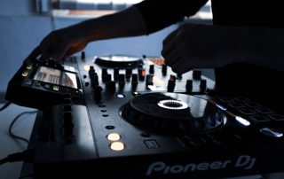 DJ controller with hands adjusting knobs and flter/turntables, glowing screens in a dim setting (Pioneer DJ brand visible)