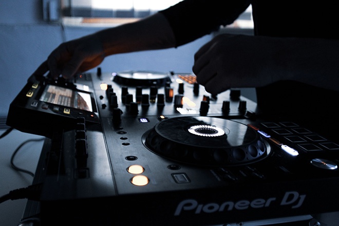 DJ controller with hands adjusting knobs and flter/turntables, glowing screens in a dim setting (Pioneer DJ brand visible)