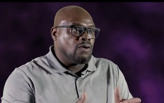 Man speaking to camera with hands raised, wearing glasses and a gray polo shirt against a purple studio background.
