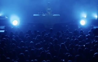 Crowd at a blue-lit concert with a DJ performing on stage in the distance.