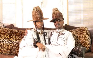 Two men in matching leopard-print outfits and tall leopard hats sit close together on a leopard-patterned couch.
