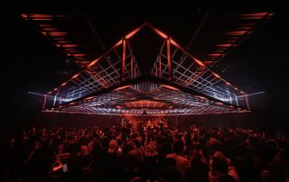 Large crowd at a dark club watching a red-lit, angular stage suspended above the dance floor.